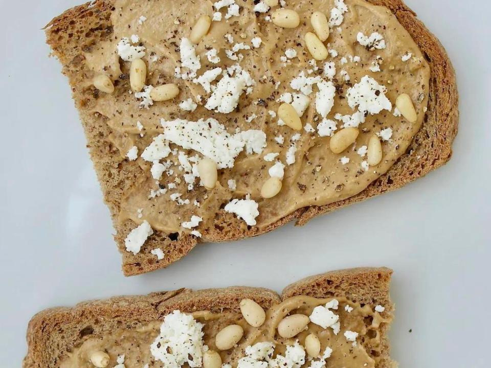 Two slices of whole-grain toast that have been topped with tahini, feta, and pine nuts.