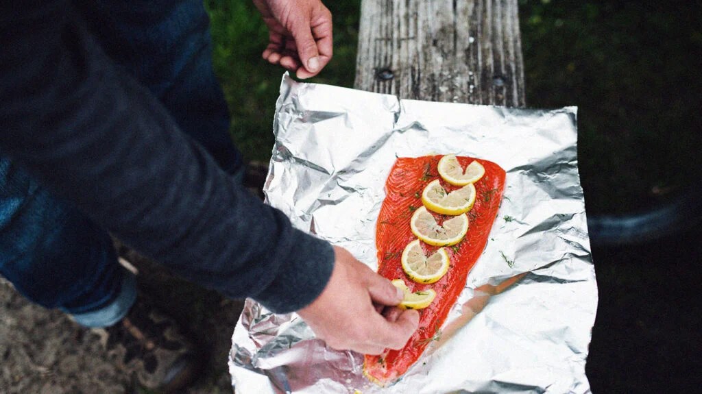 A person places lemon slices on top of a salmon fillet on a foil paper before cooking outside