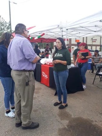 A community health worker in a green shirt, stands under a tent, speaking with two people.