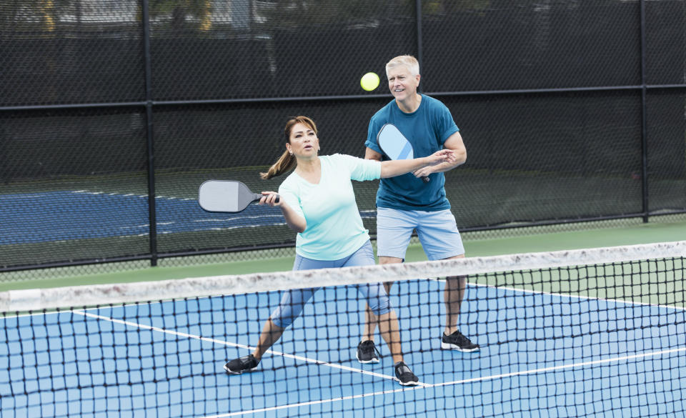 A couple playing pickleball together. They are a doubles team. The Hispanic woman, in her 50s, is concentrating on hitting the ball with her pickleball paddle. Her partner, a senior man in his 60s, is watching.